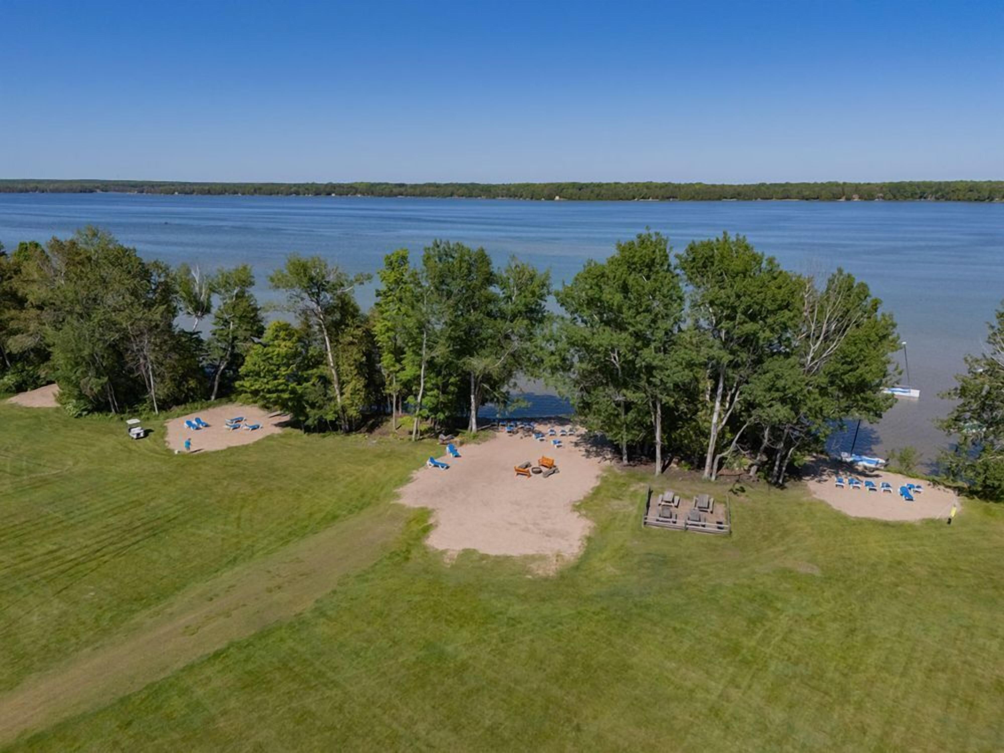 An aerial view of a lakeside property with a grassy area, several trees, and a sandy beach with lounge chairs.