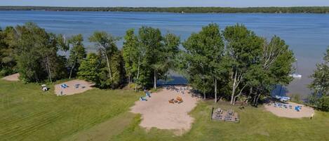 An aerial view of a lakeside property with a grassy area, several trees, and a sandy beach with lounge chairs.