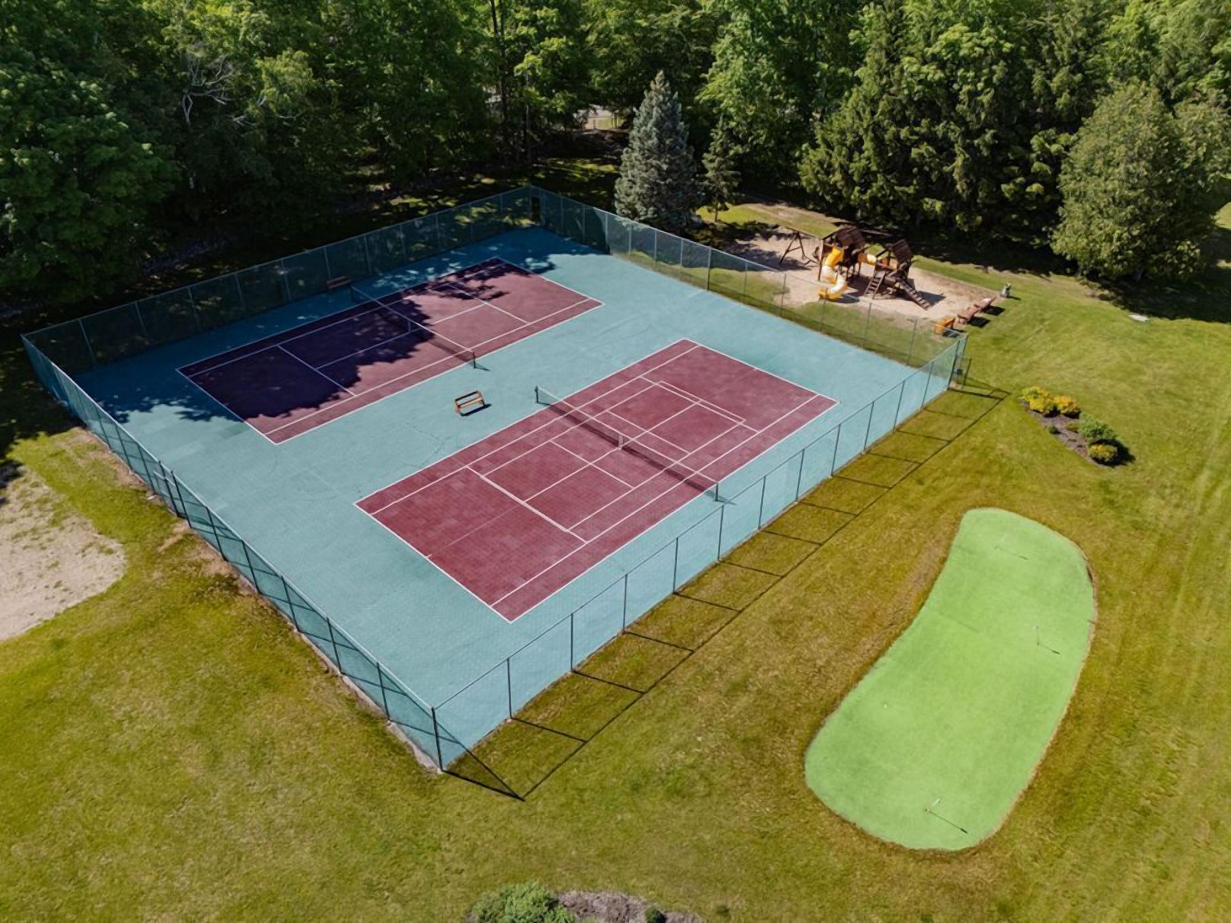 An outdoor tennis court with two courts, surrounded by a fence, and a green area for other sports.