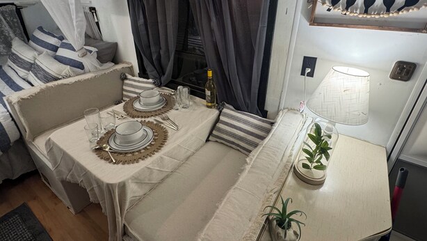 Een gezellige kamer met een eethoek, ingericht voor een maaltijd, met een tafel, borden, glazen en bestek. Er is een bank met kussens en een kleine tafel met een lamp en een bloempot.