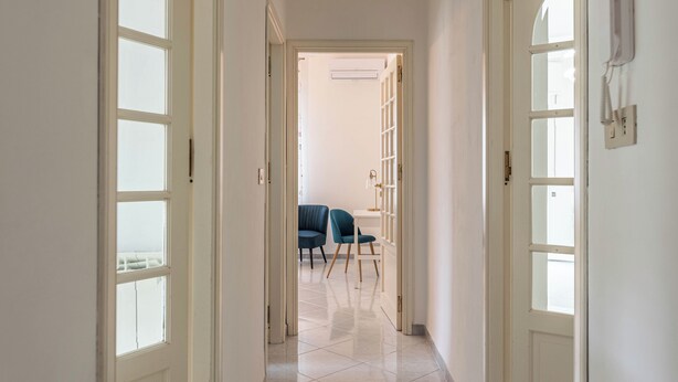 A hallway with a tiled floor, white walls, and a door leading to a room with a chair and a desk.