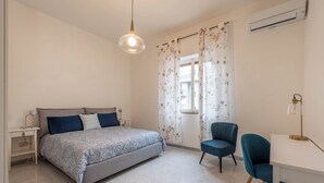 A bedroom with a bed, a desk, a chair, a nightstand, a lamp, and a window with floral curtains.