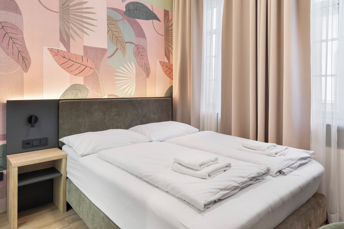 Un lit bien fait, avec du linge de lit et des serviettes blanches, une table de chevet avec une lampe, et un mur orné d’un motif de feuilles.