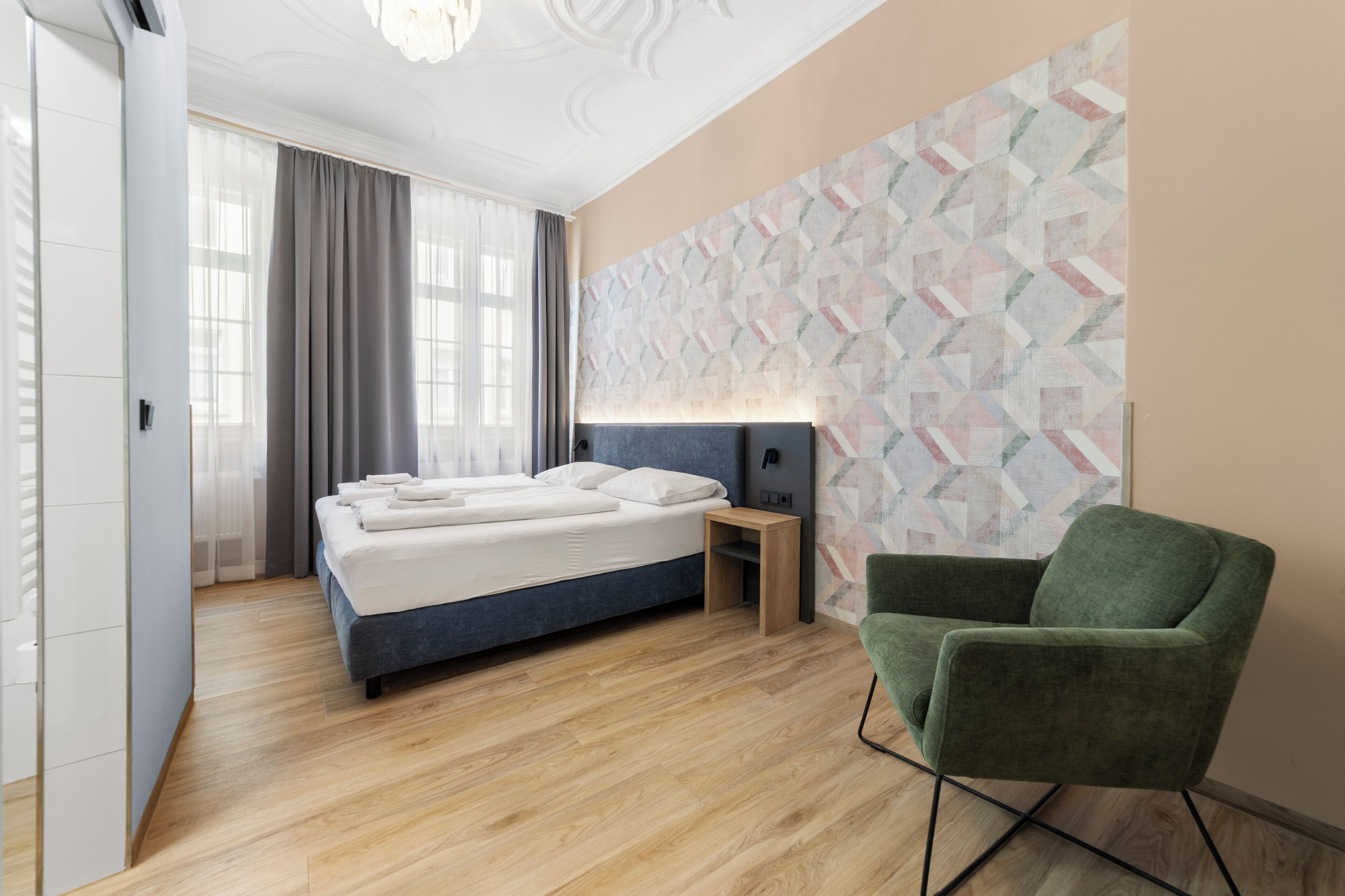 A modern hotel room with a bed, a green armchair, a nightstand, and a geometric-patterned wall.