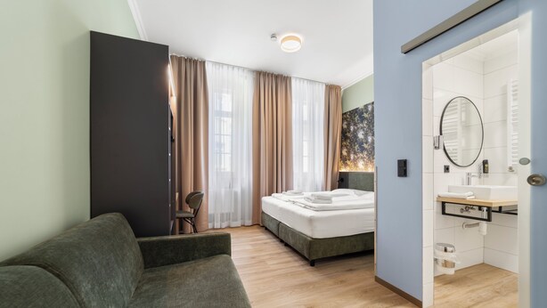 A bedroom with a bed, a chair, a bedside table, a mirror, and a bathroom with a sink and toilet. - Sona Design Rooms City Center (Bamberg)