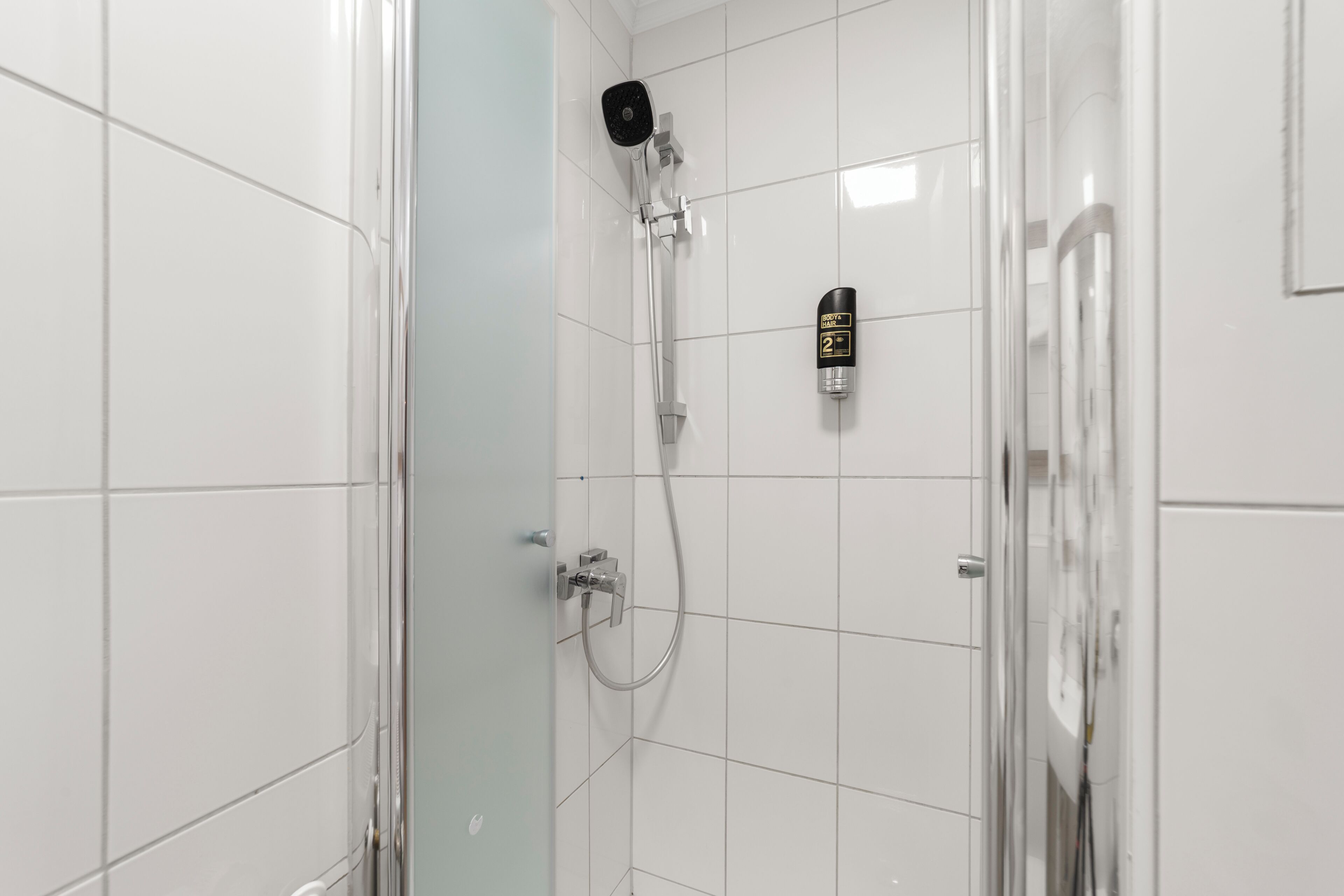 A shower with a handheld showerhead, a handheld massager, and a bottle of shampoo on the wall.