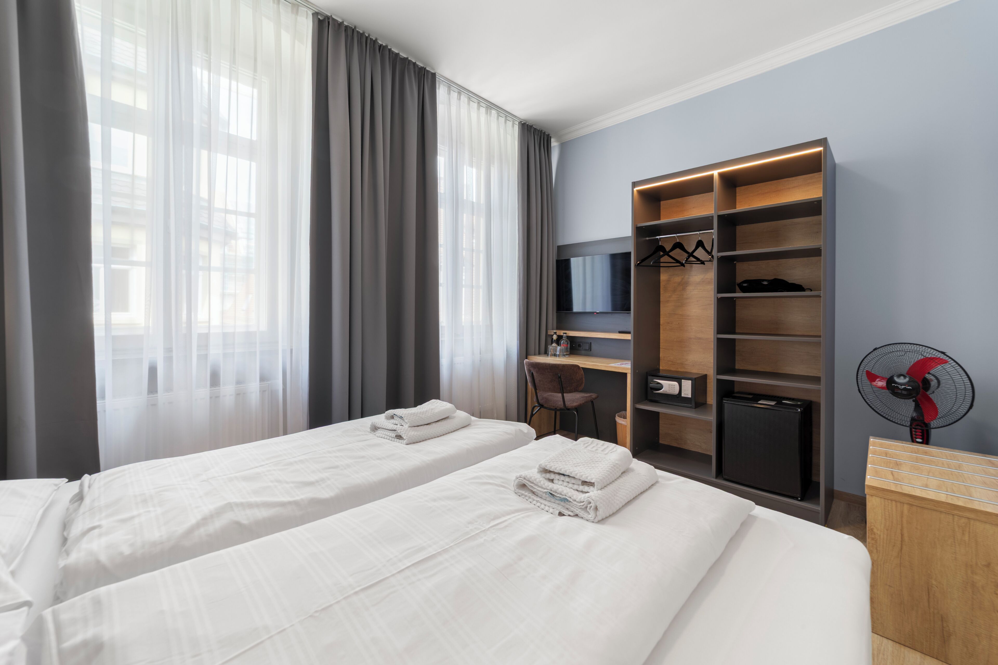 A hotel room with two beds, a desk, a chair, a TV, a wardrobe, and a fan.