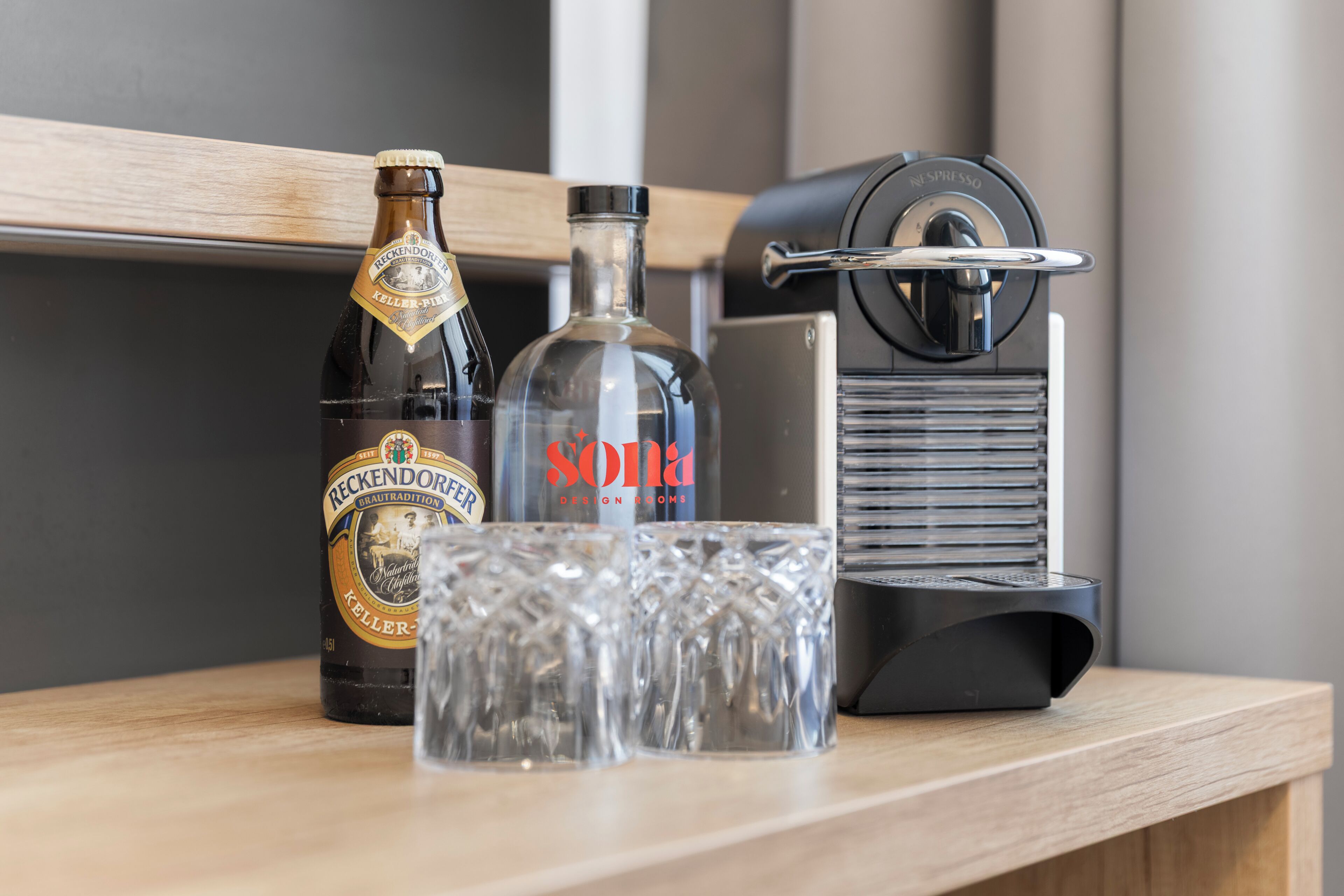 Una botella de Reckendorfer Keller-Pilsner, un vaso de Sona Design Broke y una máquina de Nespresso sobre una superficie de madera.