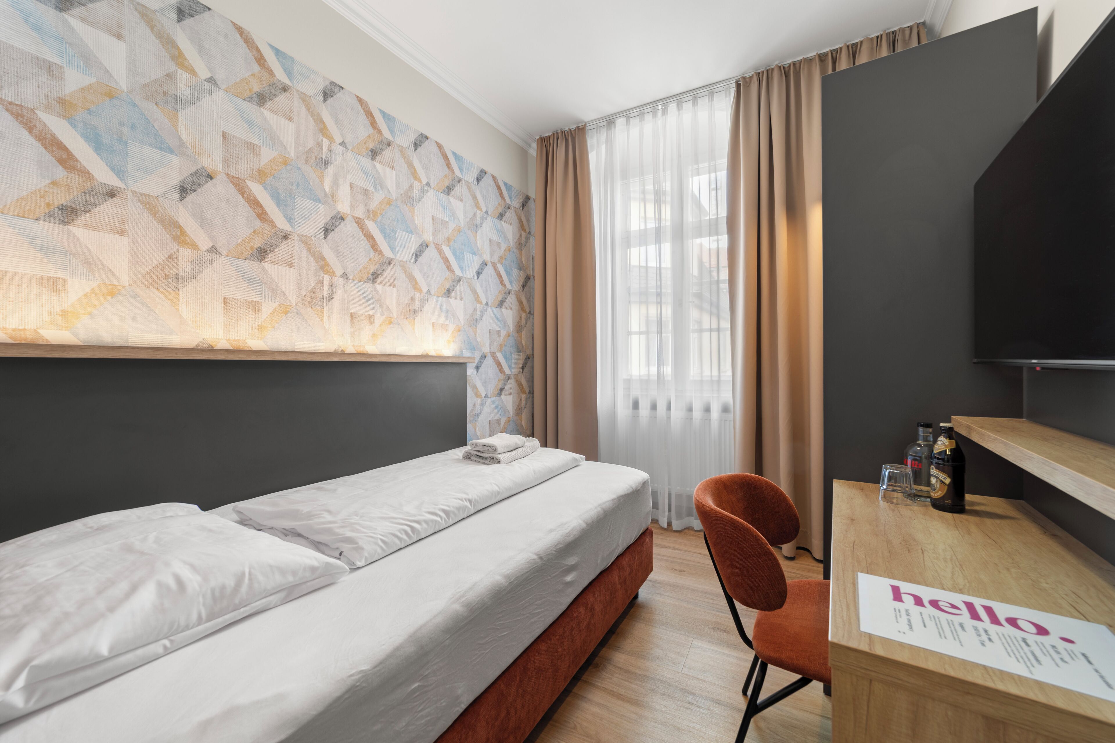 Ein Hotelzimmer mit Bett, Schreibtisch mit einem 'hello' Schild, Stuhl, Fernseher und einer Wand mit Muster.
