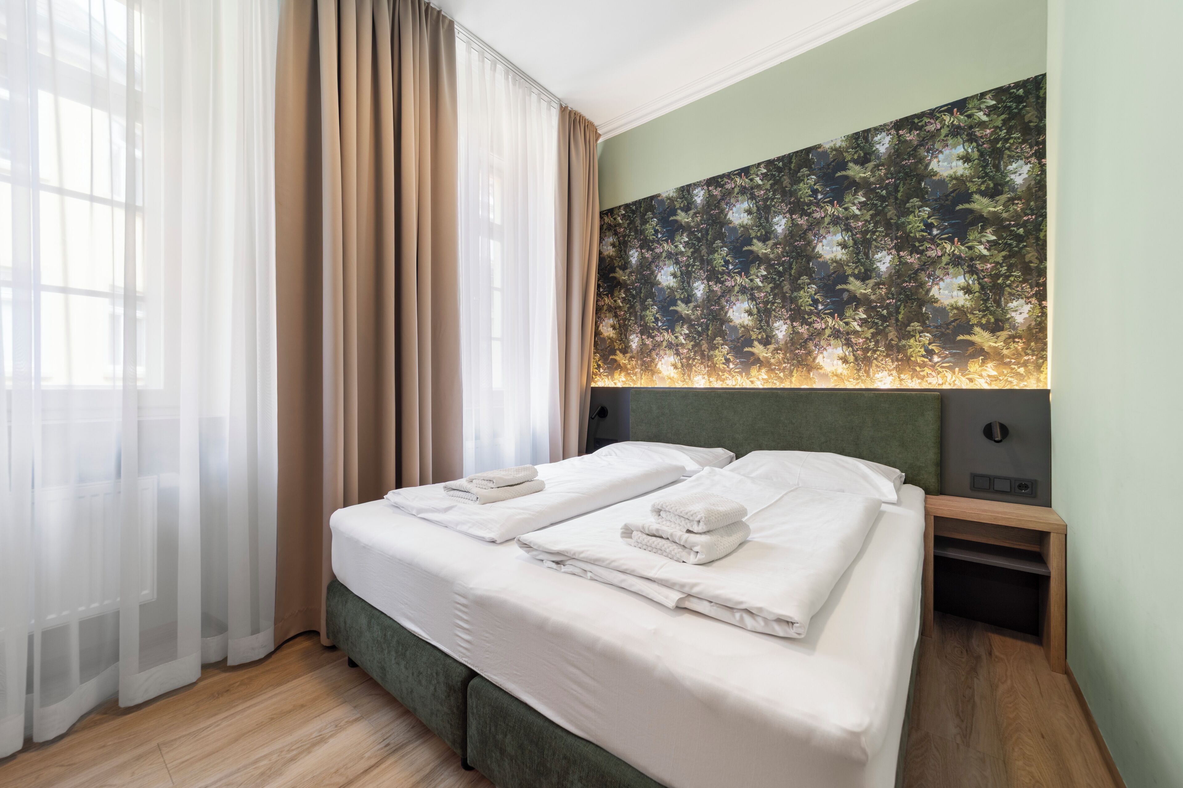 Una habitación de hotel con cama, mesita de noche, televisor montado en la pared y cortinas traslúcidas.