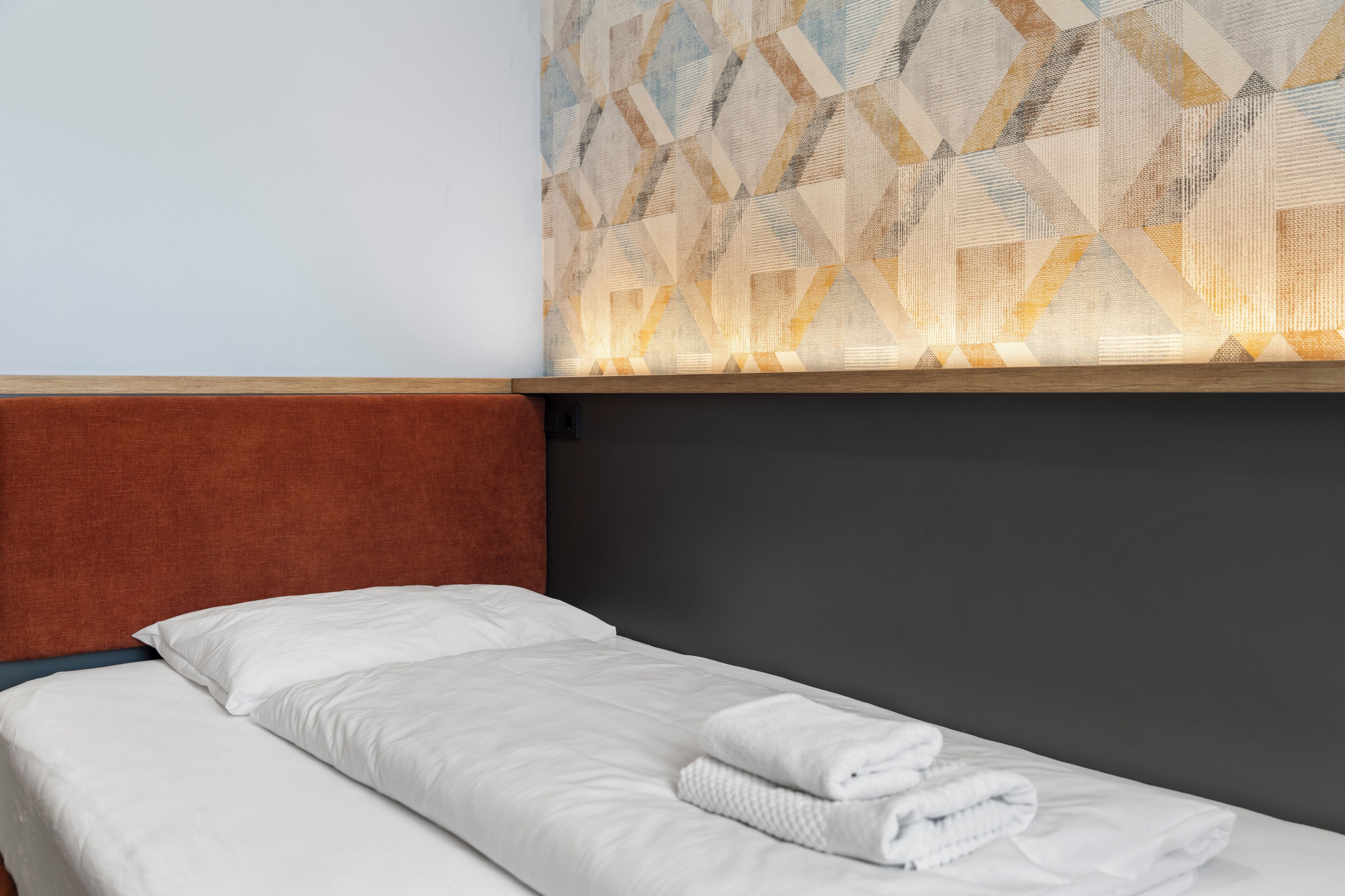 Una cama individual con ropa blanca, cabecera marrón y un papel tapiz estampado en la pared.
