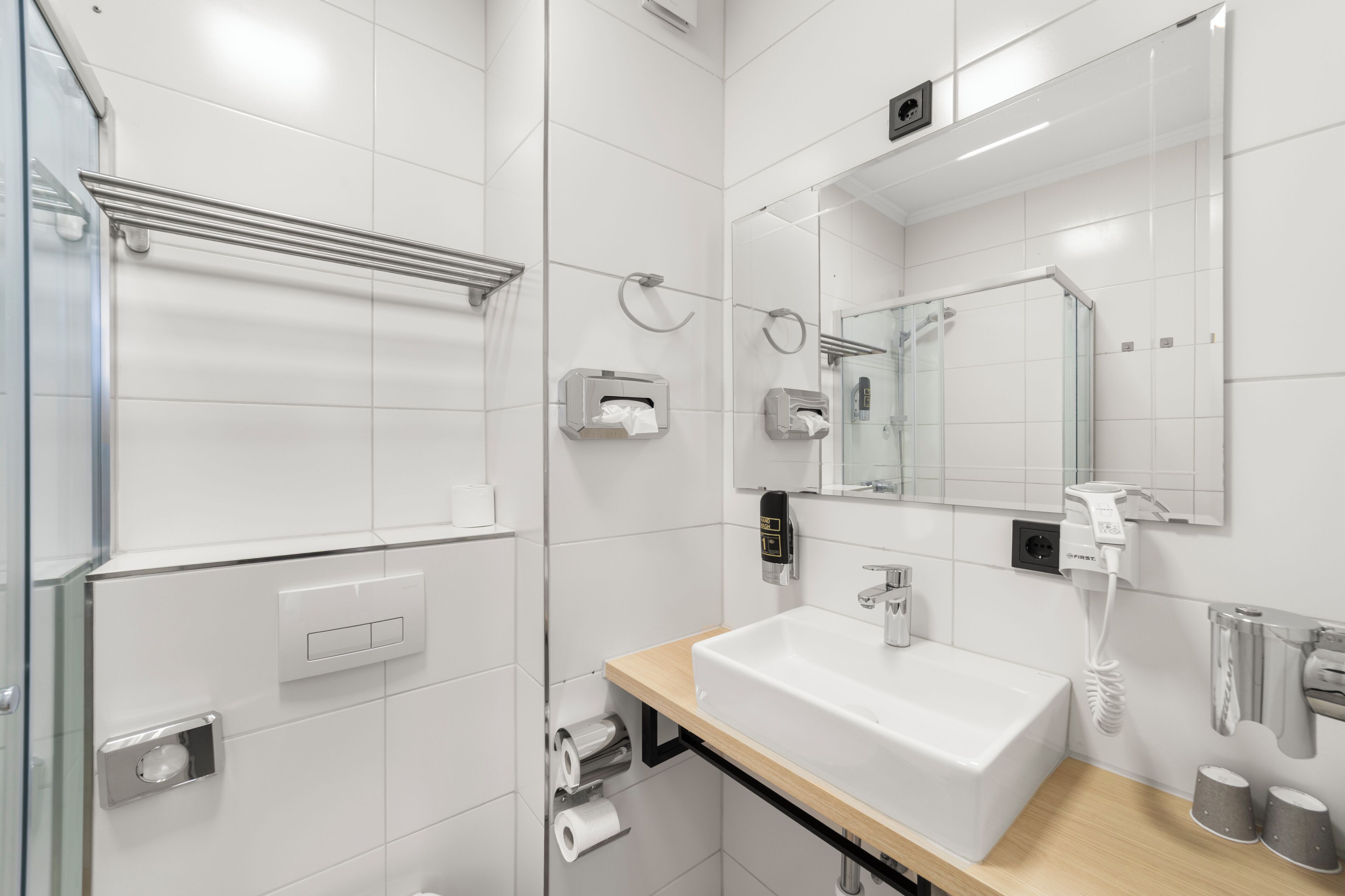 A modern bathroom with a white sink, mirror, towel dispenser, and toilet paper.