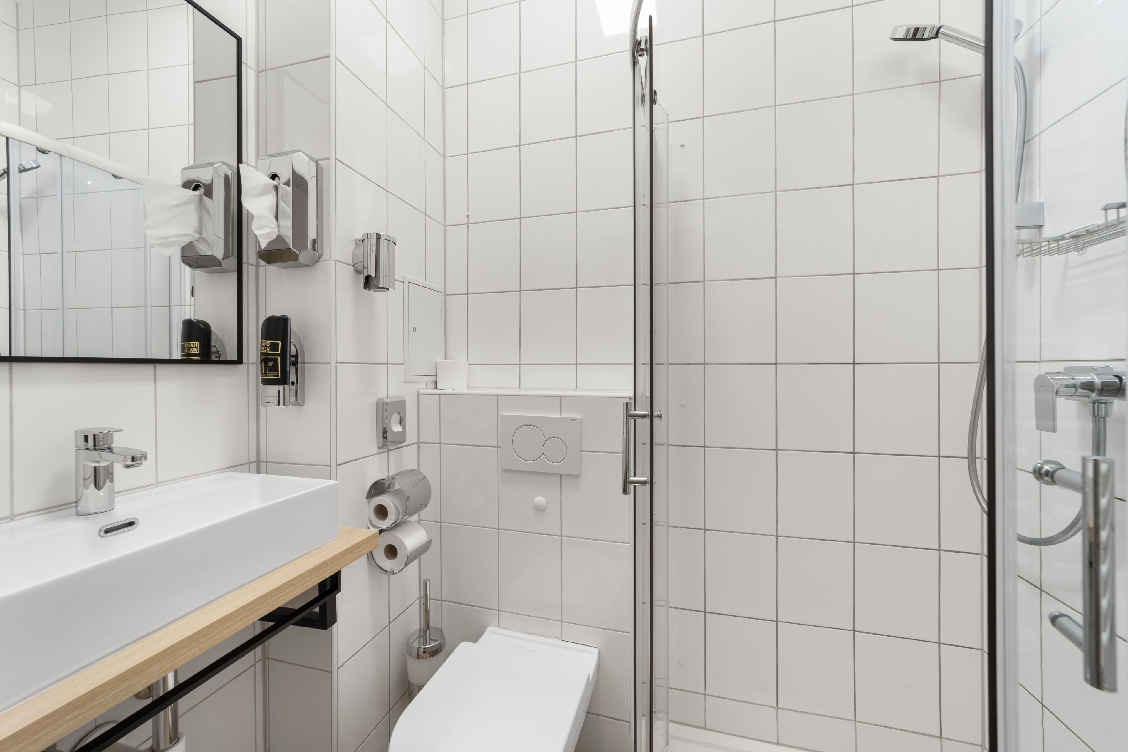 Une salle de bain moderne avec un lavabo blanc, un miroir, un distributeur de serviettes, du papier toilette et une cabine de douche en verre.