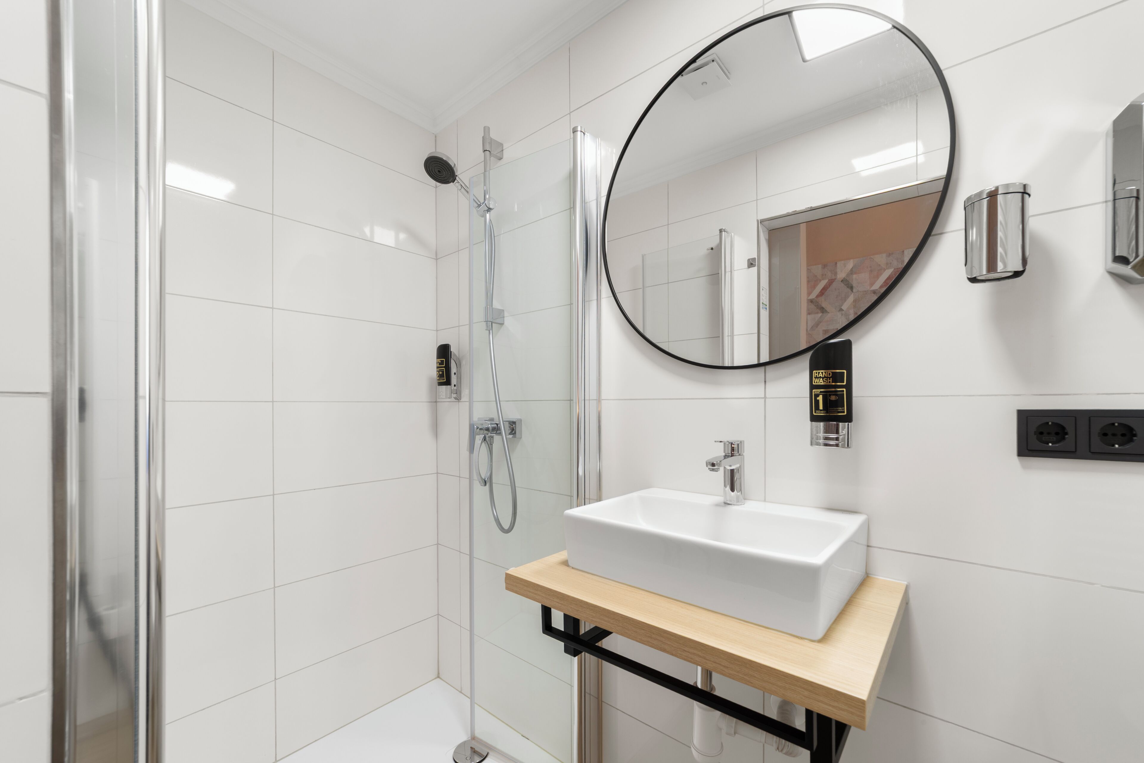 Un baño moderno con cabina de ducha de vidrio, un lavamanos blanco y encimera de madera, y un espejo redondo.