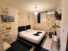 Una habitación pequeña con paredes de piedra, una cama, un cuadro, una lámpara colgante y un tendedero.