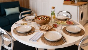 A dining table set with plates, bowls, glasses, and bread, surrounded by chairs in a modern kitchen and living area.