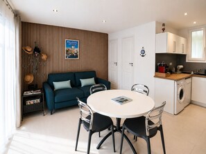Een moderne keuken met een wasmachine, een eethoek met een ronde tafel en stoelen, een blauwe bank en een houten wand met een ingelijst kunstwerk.