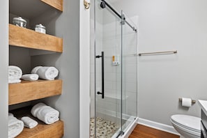 A bathroom with a glass shower enclosure, wooden shelves with towels, and a toilet.