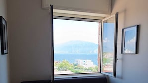 A room with a dark-colored sofa, a dining table, and a large window offering a view of buildings and mountains. - Torre del Greco waterfront apartment - 6 floor with panoramic view  (Torre del Greco)
