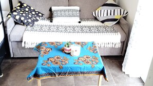 Un salon avec un canapé gris, des coussins à motifs, une table bleue ornée d’une plante et d’une bougie, et un mur blanc décoré d’un motif circulaire.