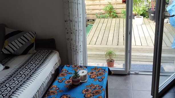Une pièce chaleureuse avec un canapé et une table, et une porte-fenêtre coulissante donnant sur une terrasse en bois agrémentée de plantes.