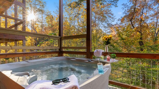 A hot tub with a towel and sunglasses, surrounded by a wooden deck and a glass railing, overlooking a forest with autumn foliage. - Elegant Elite Estate By Ghosal Luxury Lodging (Pigeon Forge)