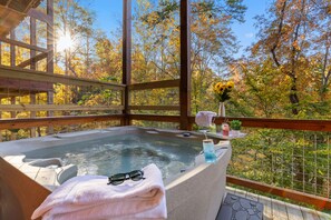 A hot tub with a towel and sunglasses, surrounded by a wooden deck and a glass railing, overlooking a forest with autumn foliage. - Elegant Elite Estate By Ghosal Luxury Lodging (Pigeon Forge)