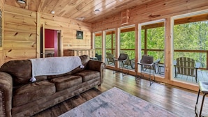 A wooden cabin interior with a brown leather couch, a red wall, and a large window overlooking greenery. - Elegant Elite Estate By Ghosal Luxury Lodging (Pigeon Forge)