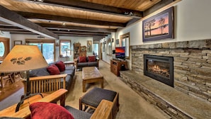 A cozy living room with a stone fireplace, wooden ceiling beams, and comfortable seating. - Spacious West Shore Cabin! (Homewood)