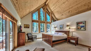 A bedroom with a large bed, wooden ceiling, and large windows offering a view of trees. - Spacious West Shore Cabin! (Homewood)