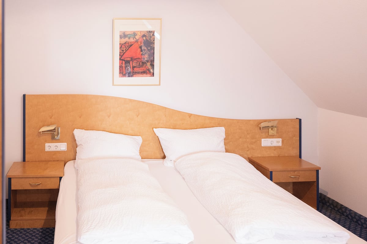 Una cama doble con ropa blanca, dos mesitas de noche con lámparas, un cuadro en la pared y un cajón pequeño debajo de la cama.