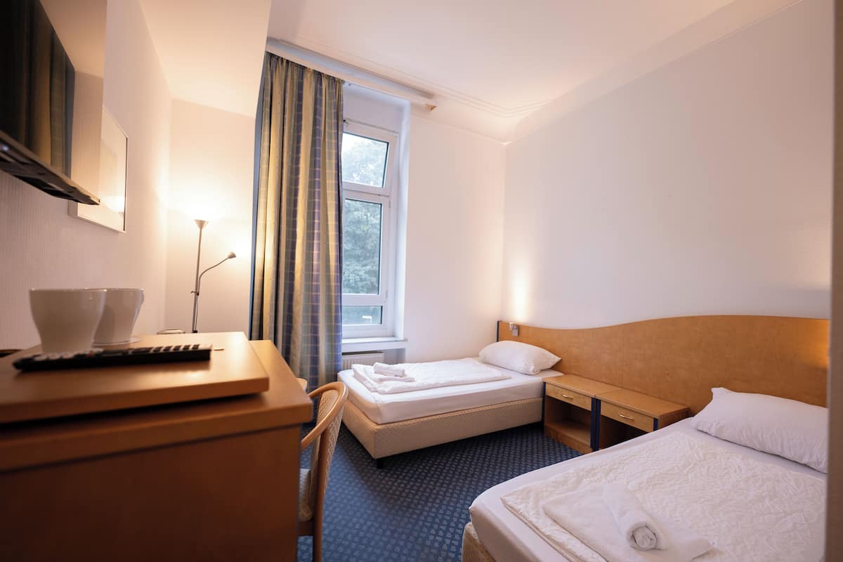 Habitación de hotel con cama, escritorio, silla, mesita de noche, lámpara, ventana con cortinas y control remoto.