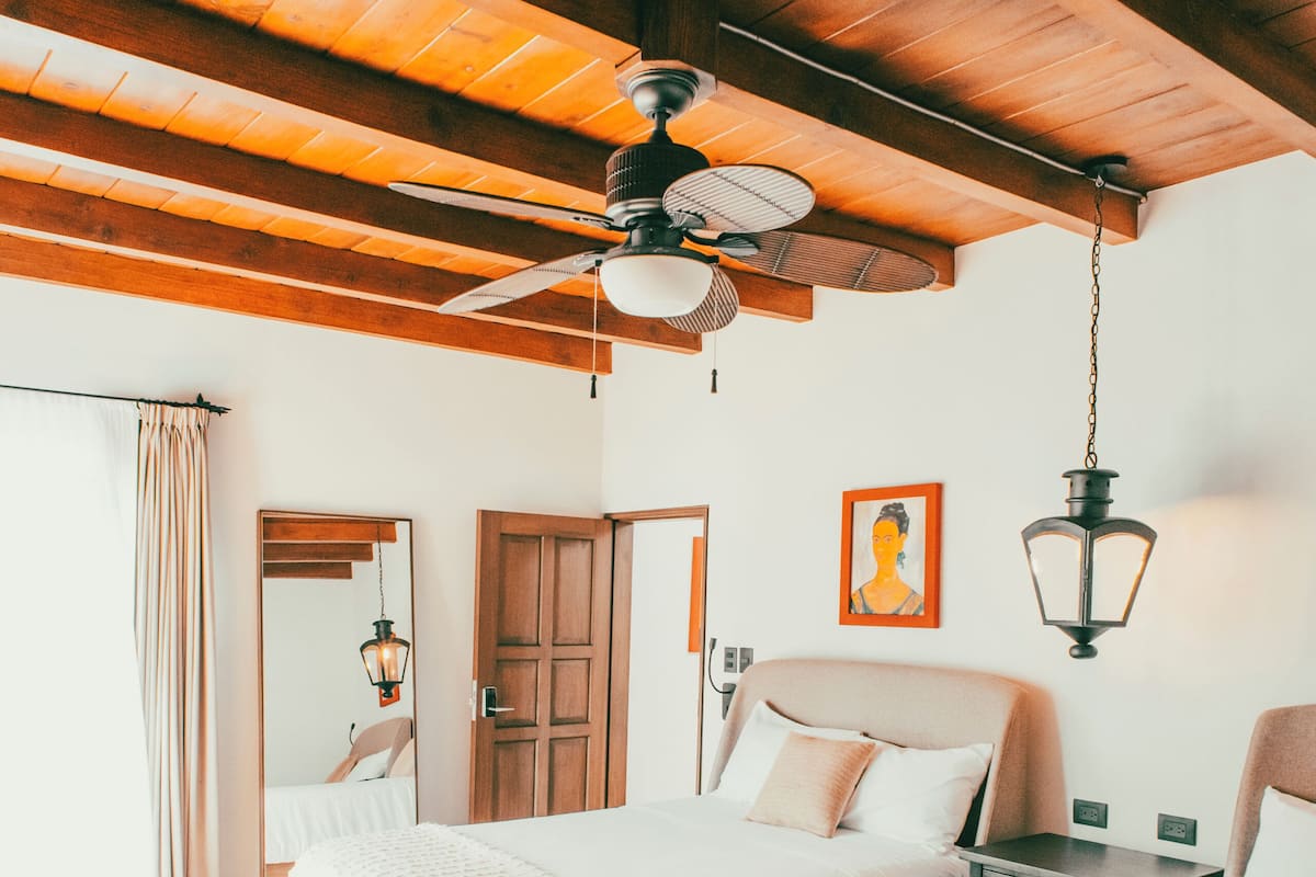 Quarto com duas camas, vigas de madeira no teto, um ventilador de teto, um candeeiro pendente, um banco e uma porta.