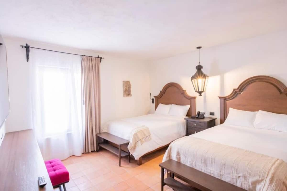 Quarto de hotel com duas camas, um banco, uma escrivaninha e um abajur.