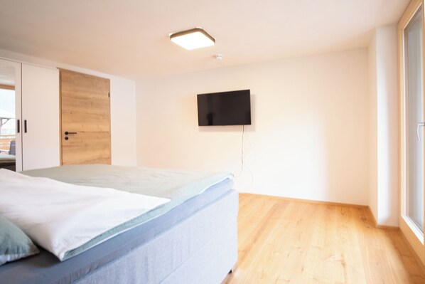 A bedroom with a bed, a wooden wardrobe, a television mounted on the wall, and a large window.