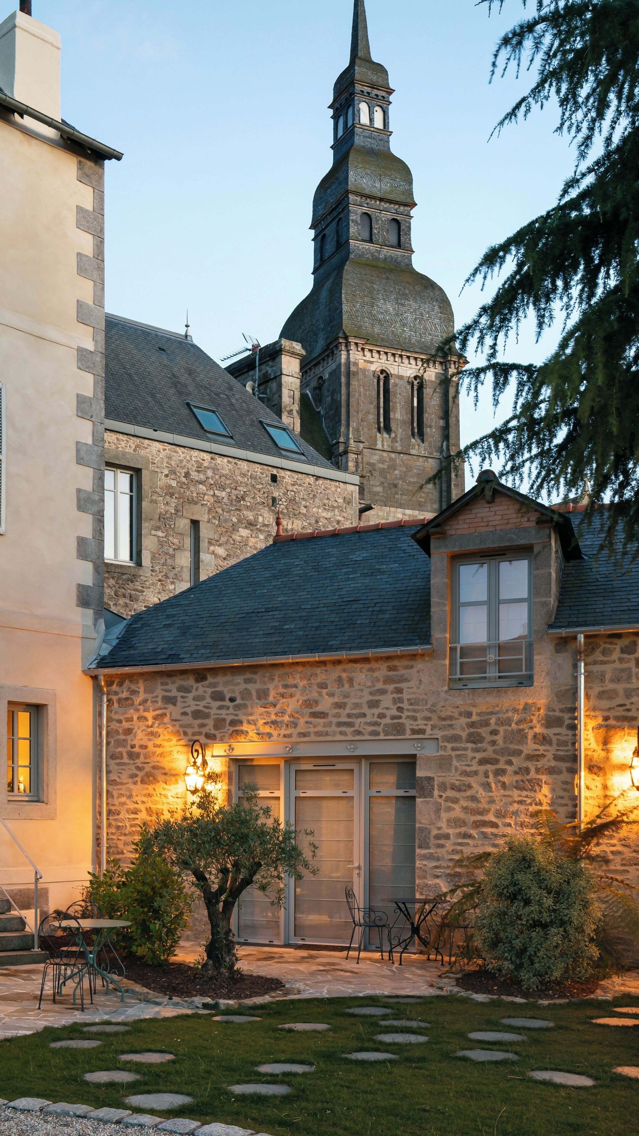 Un bâtiment en pierre avec un toit en pente, une tour surmontée d’une flèche et un petit jardin agrémenté de pierres plates servant de pas.