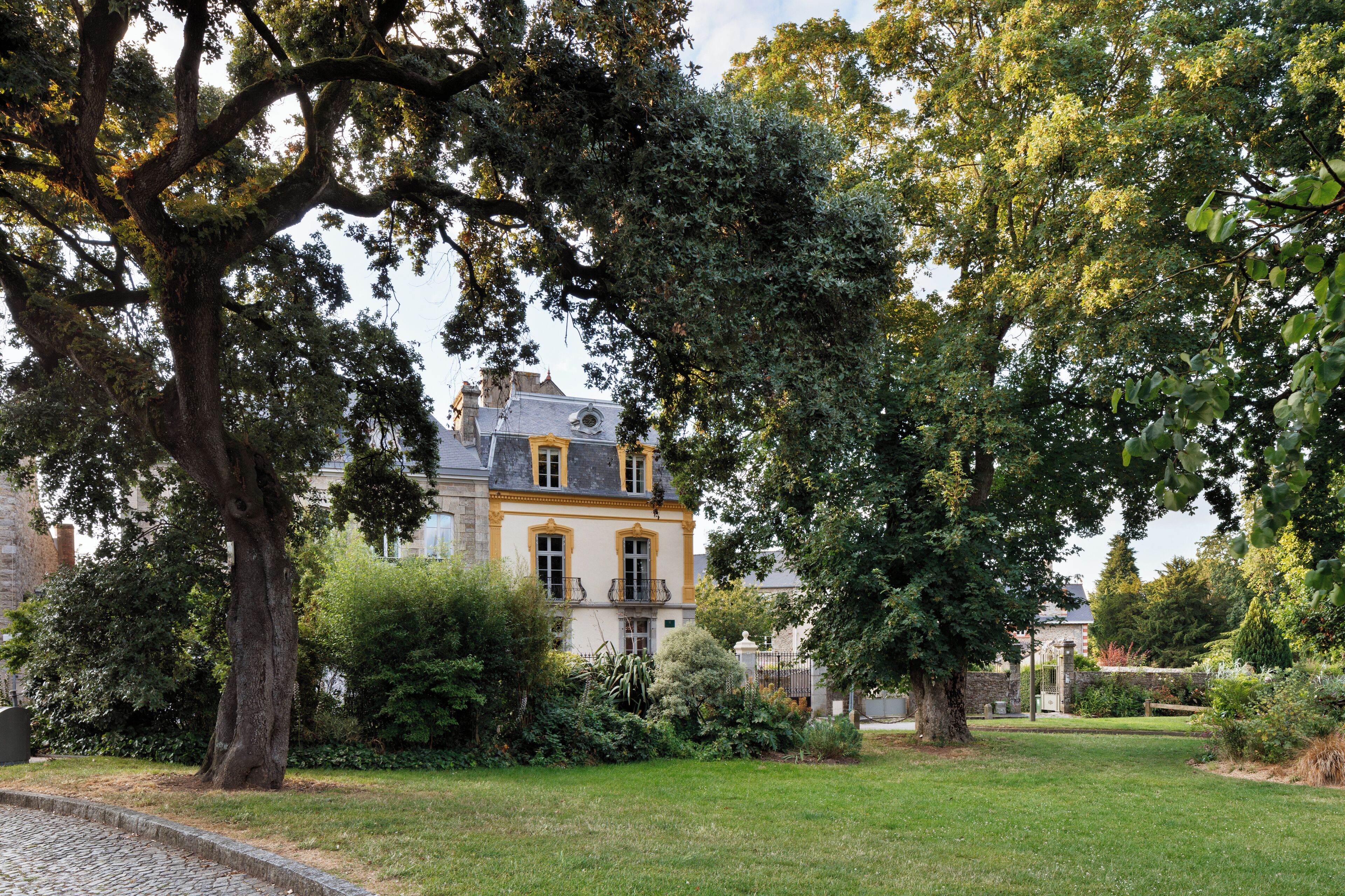 A historic building with a large tree in the foreground, surrounded by well-maintained gardens and a cobblestone path.