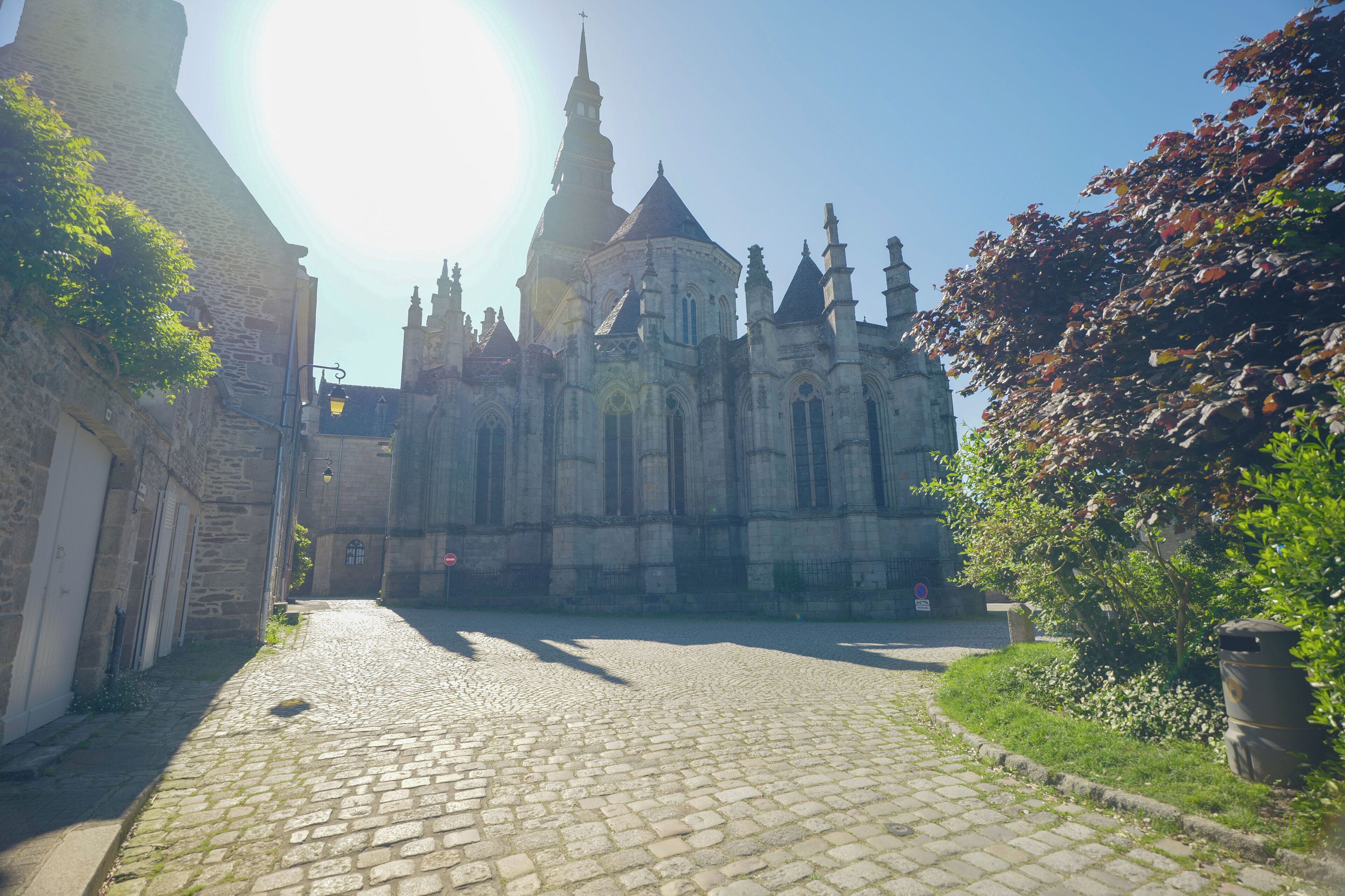 Ruas de paralelepípedos que levam a uma catedral histórica com uma torre alta, rodeada por edifícios de pedra e vegetação.