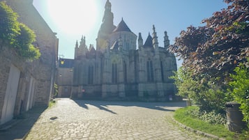 Ruas de paralelepípedos que levam a uma catedral histórica com uma torre alta, rodeada por edifícios de pedra e vegetação.
