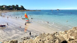 A beach with a sailboat, people swimming, and a castle in the background. - Clos du Cèdre (Dinan)