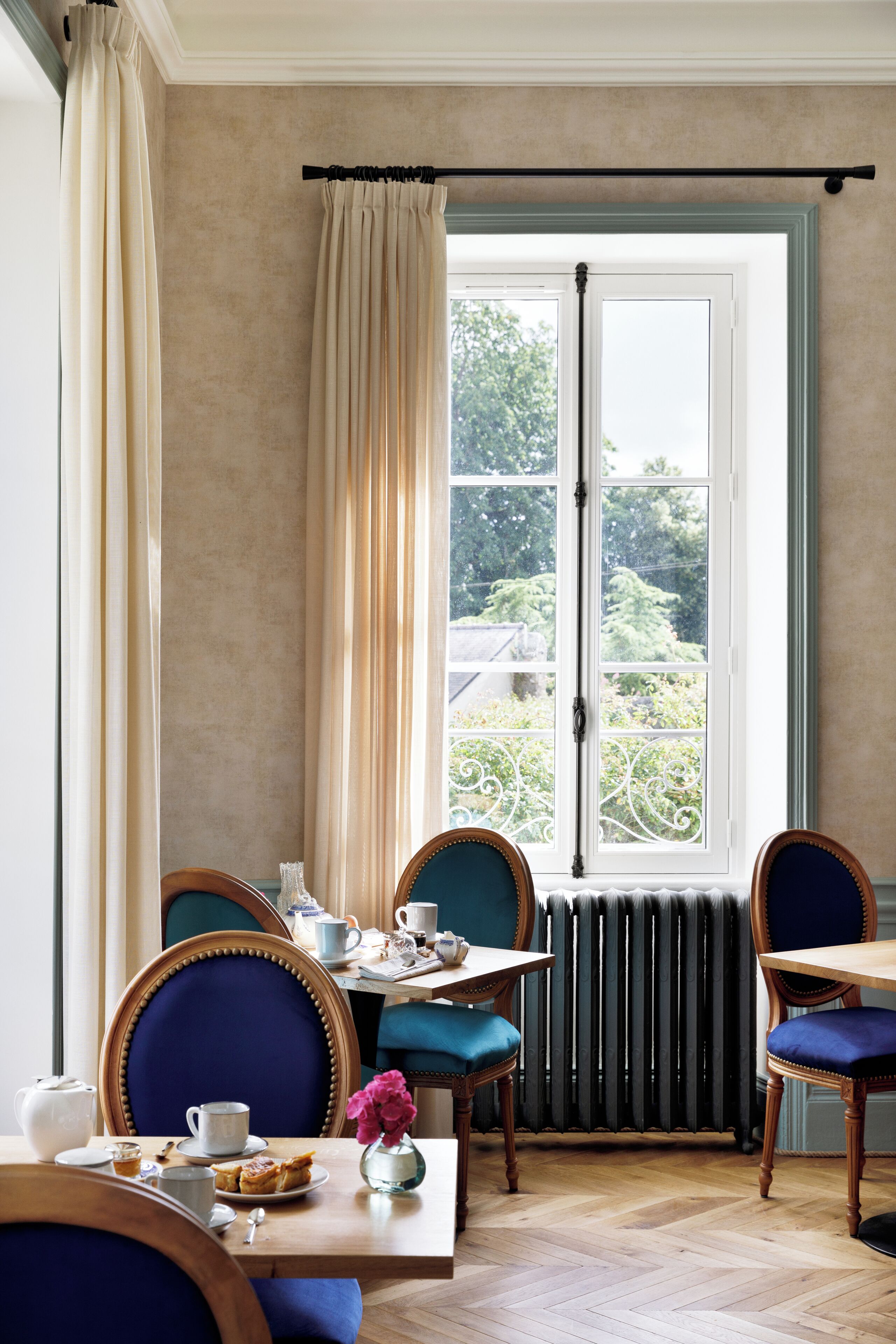 Un coin repas avec une table dressée pour deux personnes, des chaises recouvertes d’un tissu bleu, une fenêtre avec des rideaux et un radiateur en dessous.