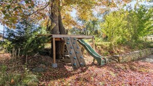 A wooden climbing structure with a slide, surrounded by trees and autumn leaves. - TreeFort, ZipLine, Kayaks, Beach6min drive, Vaulted Ceilings, ScreenPorch & Deck (Narragansett)