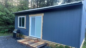 A small, single-story building with a dark exterior, a wooden deck, and a small outdoor grill. - Cozy Cabin with Stunning Views (Seward)