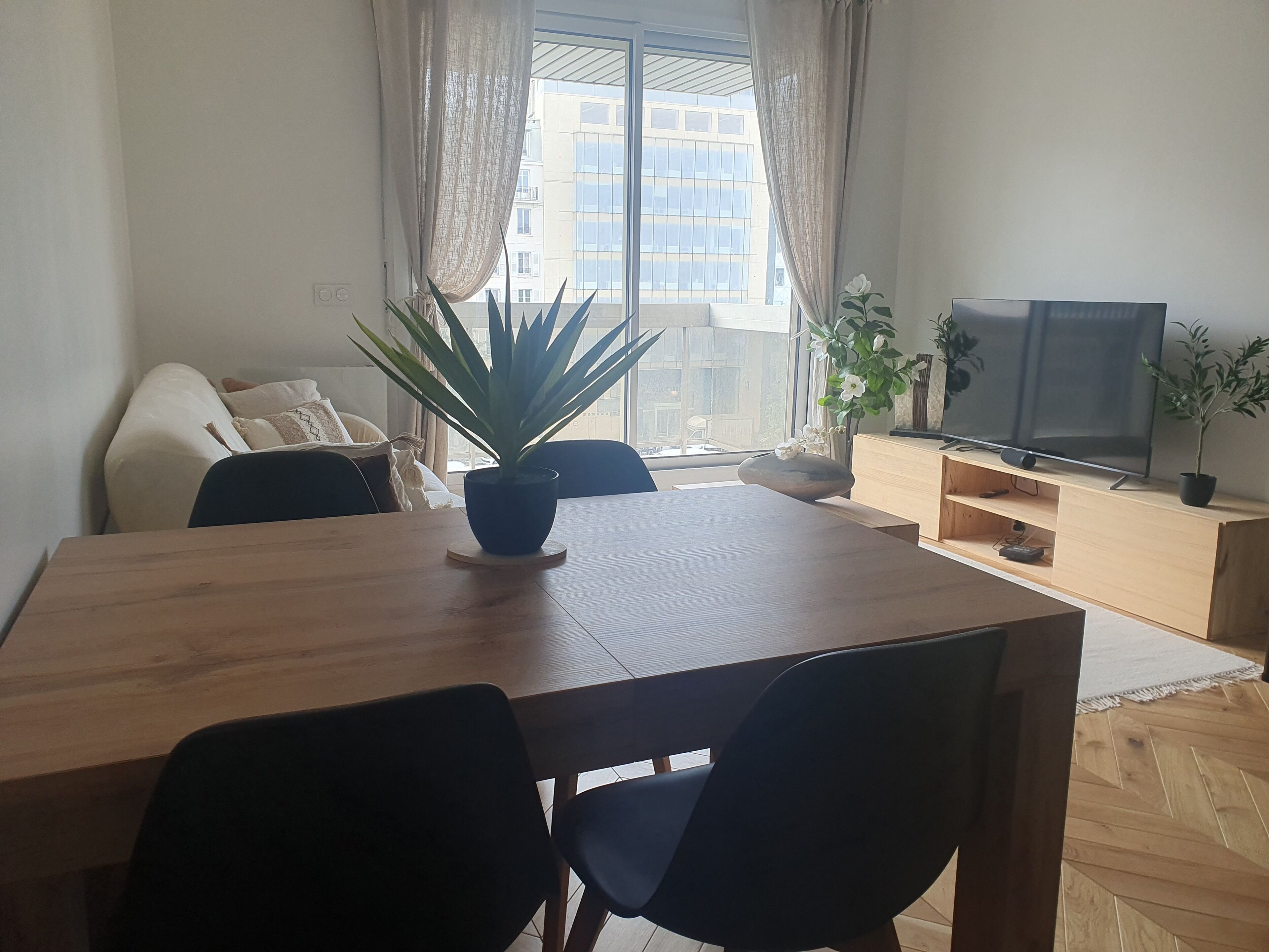 Uma sala de estar moderna com uma mesa de jantar de madeira, cadeiras pretas, uma televisão e plantas em vasos.