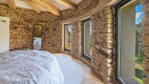 A bedroom with stone walls, a wooden ceiling, and large windows.