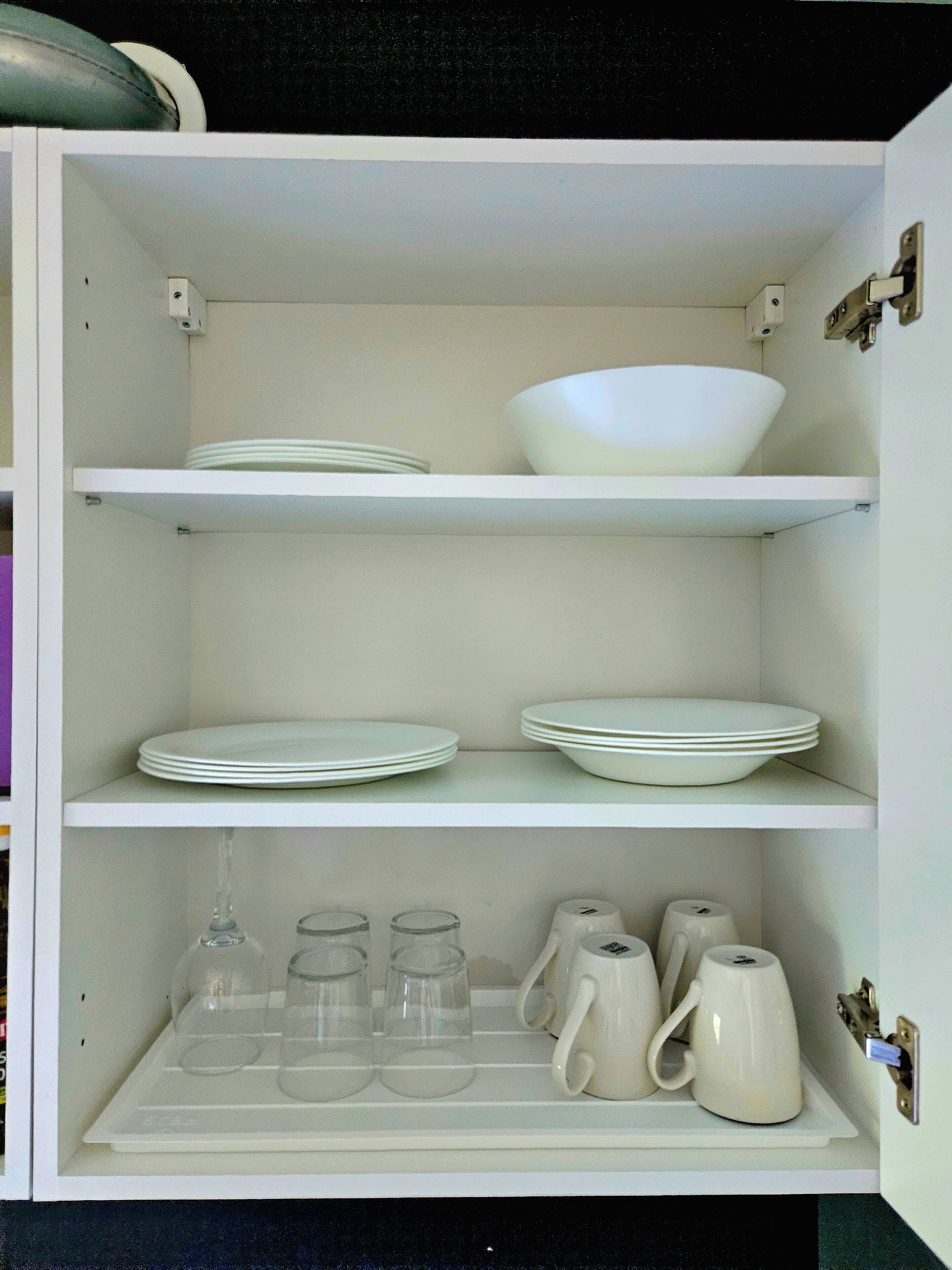 A white cabinet with shelves containing white plates, bowls, glasses, and a teapot.