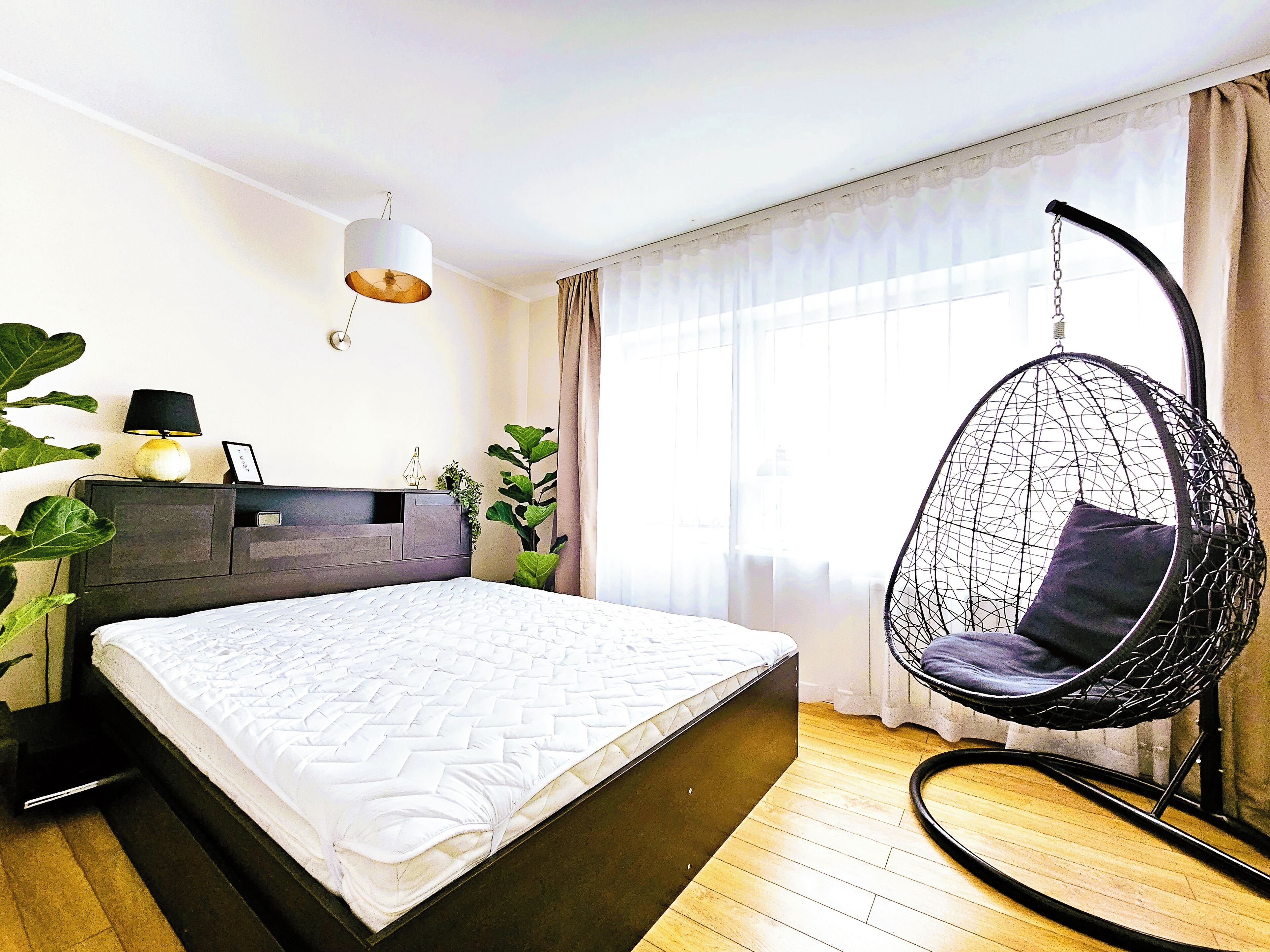 A modern bedroom with a hanging chair, a bed, a bedside table with a lamp, and a large window with sheer curtains.