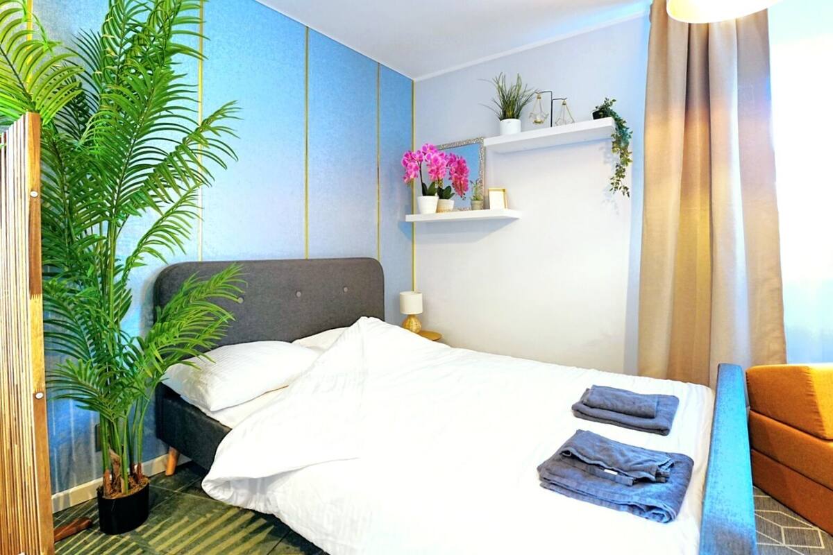 A neatly made bed with white linens, a grey headboard, and a potted plant beside it.