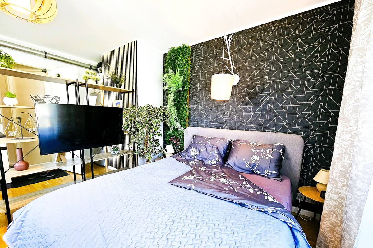 A modern bedroom with a large bed, a flat-screen TV, a geometric-patterned wall, and a green wall with plants.