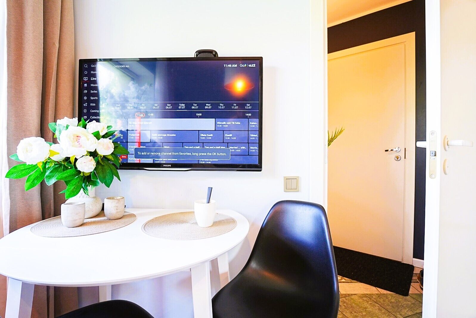 Habitación de hotel con un televisor de pantalla plana montado en la pared mostrando una guía de televisión. Hay una mesa de comedor con un jarrón de flores y algunas tazas. Una silla negra está ubicada frente a la mesa.
